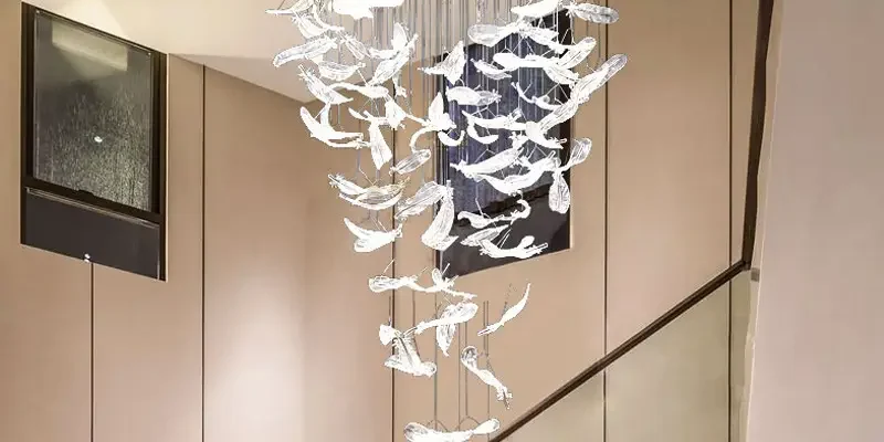 Intricate glass feather chandelier with cascading translucent feathers hangs in a modern space, creating an artistic, ambient glow against a blue-toned window background.