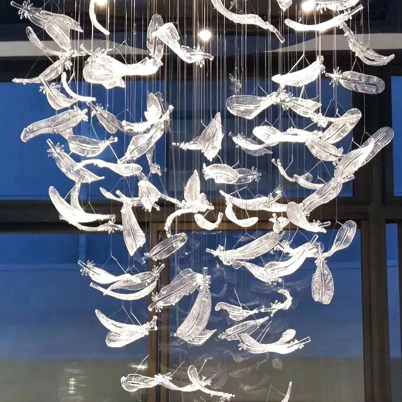 Intricate glass feather chandelier with cascading translucent feathers hangs in a modern space, creating an artistic, ambient glow against a blue-toned window background.