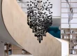 Black Stone Charcoal Chandelier suspended in a modern library or luxury retail atrium. The black stone (or charcoal) sculptural light fixture hangs dramatically within a curved wooden staircase well, set against tall built-in bookshelves and abstract ink-wash wall panels.