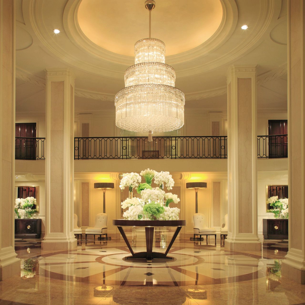 A grand hotel lobby with a tiered crystal chandelier, tall columns, and a large floral arrangement at its center.